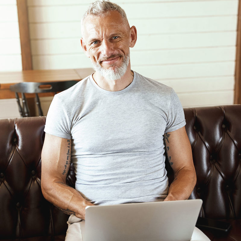Home office. Portrait of a happy middle aged man in casual clothes using laptop and smiling at camera while sitting on sofa in the living room. Freelance or distance work, self isolation concept