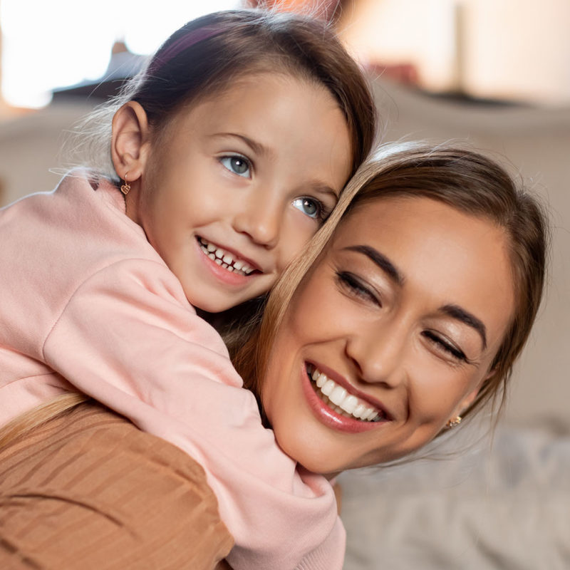 Satisfied pretty woman and little girl playing and spending time together in living room interior. Cute daughter embracing mommy from behind riding on her back. Mother's day celebration